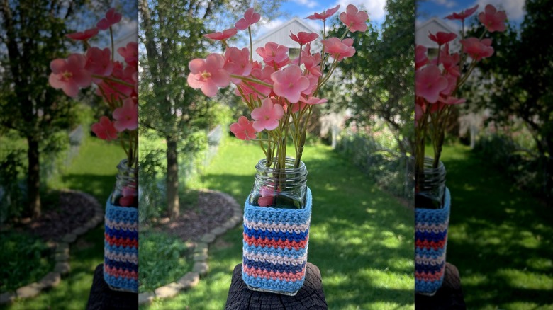 A spice jar with a crochet cover and flowers inside sits on a wooden post in front of a garden.