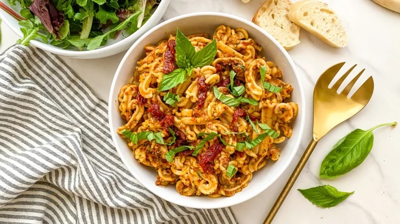 Sun-dried tomato pesto pasta in a bowl with basil leaves, bread, and a salad