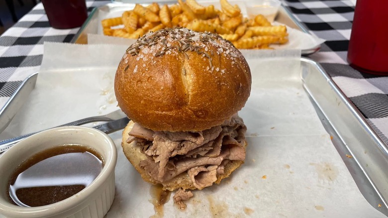 A beef on weck sandwich on a tin tray with a side of dipping sauce and fries in the background, on a plaid tablecloth
