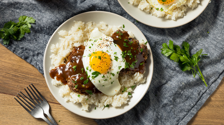 A plate of Hawaiian Loco Moco on a wooden table with a gray tablecloth, with another plate and forks partially in view