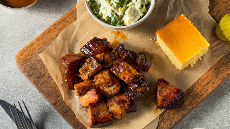 Burnt ends on parchment paper on a wooden cutting board with a side and cornbread, on a counter with a fork and knife