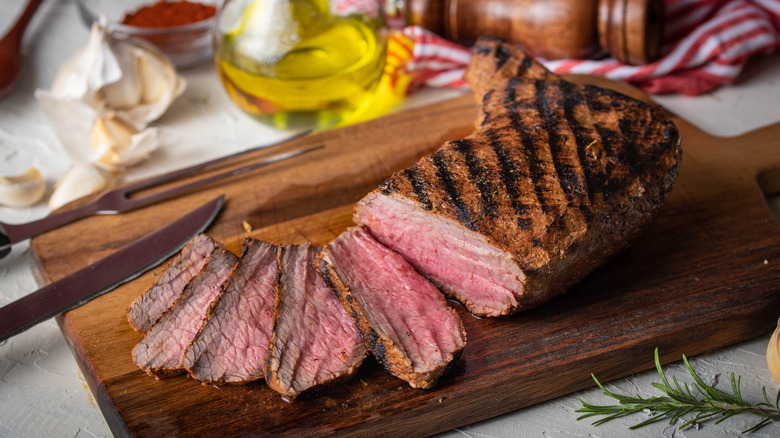 A half-sliced Santa Maria tri-tip steak on a wooden cutting board on a kitchen counter with garlic, spices, and oil in the background