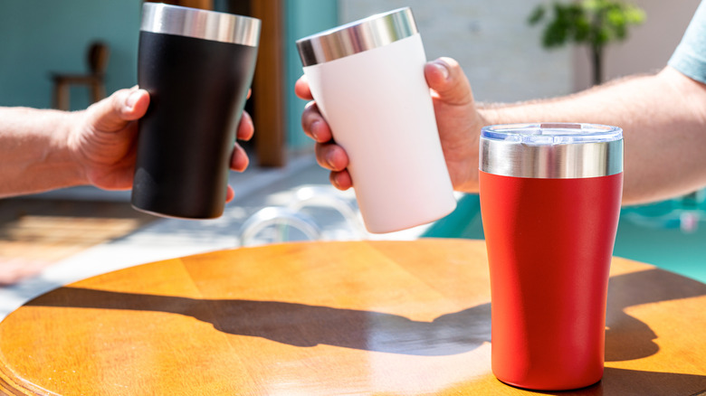 Hands holding insulated black and white tumblers with a red insulated tumbler on a table