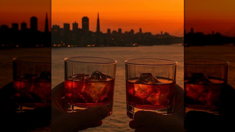 Two hands holding two glasses of whiskey on ice at sunset with the San Francisco skyline in the background.