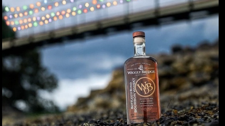 A bottle of bourbon on a rocky coast with a lit up suspension bridge in the background.