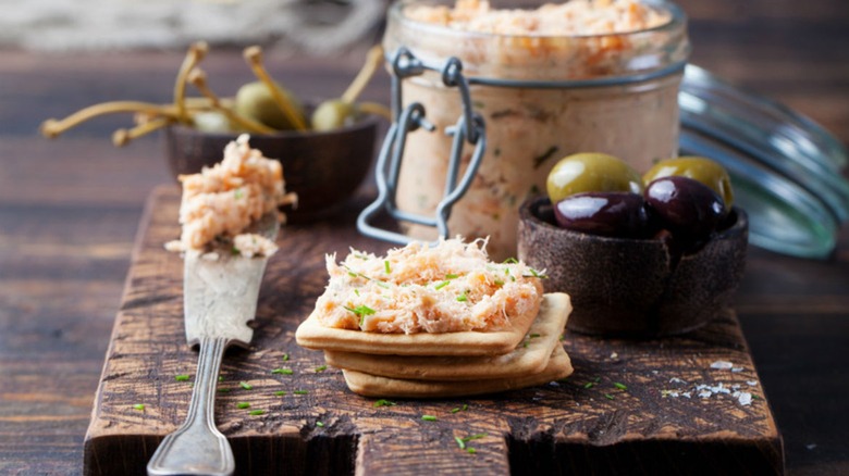 Creamy smoked salmon mousse in a jar and on crackers served on a wooden cutting board with olives