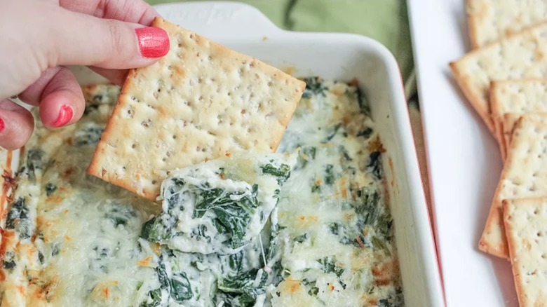 Hand dipping crackers into hot creamy spinach dip in casserole dish