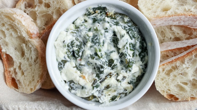 Creamy spinach dip in white bowl surrounded by freshly-cut bread slices