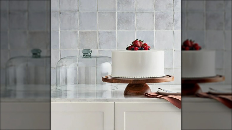 Wooden cake stand with white fruit-topped cake on countertop with cloth napkins