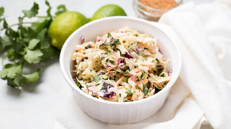 Creamy slaw in white bowl next to bunch of cilantro and fresh limes