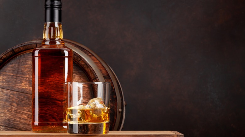 A composite image of an unbranded whiskey bottle and a full glass in front of a barrel