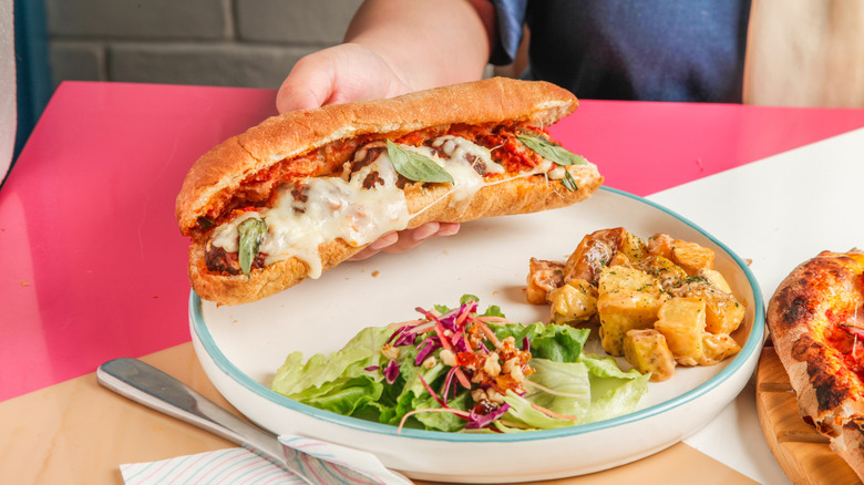 Hand holding a meatball sub over a plate of salads while sitting at a table