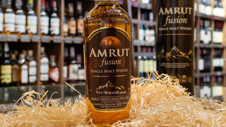 A bottle of Amrut Fusion posed among straw in a liquor store with its box shown just behind it