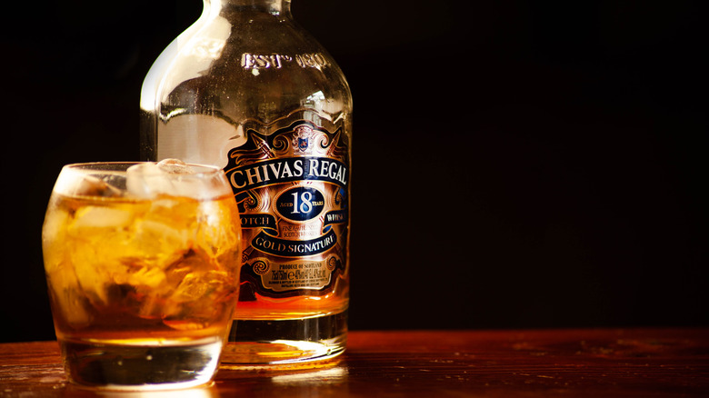 A bottle of Chivas Regal 18-year next to a filled glass in front of a black background