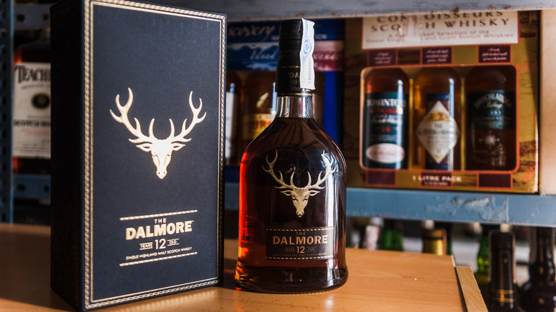 A bottle of Dalmore 12-year on a table next to its box with other whisky bottles in the background