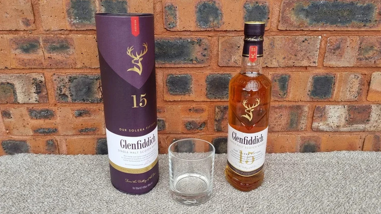 A bottle of Glenfiddich 15-year next to a branded glass and a box with a red brick background