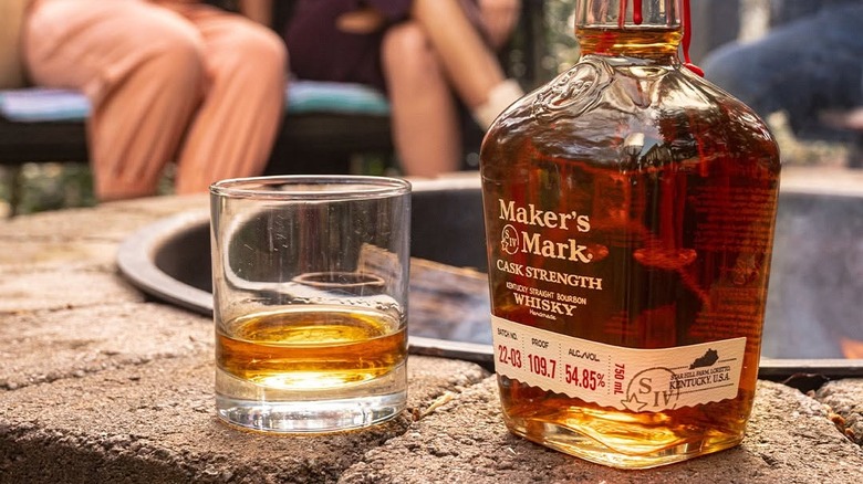 A bottle of Maker's Mark Cask Strength placed on the rim of a fire pit next to a filled glass