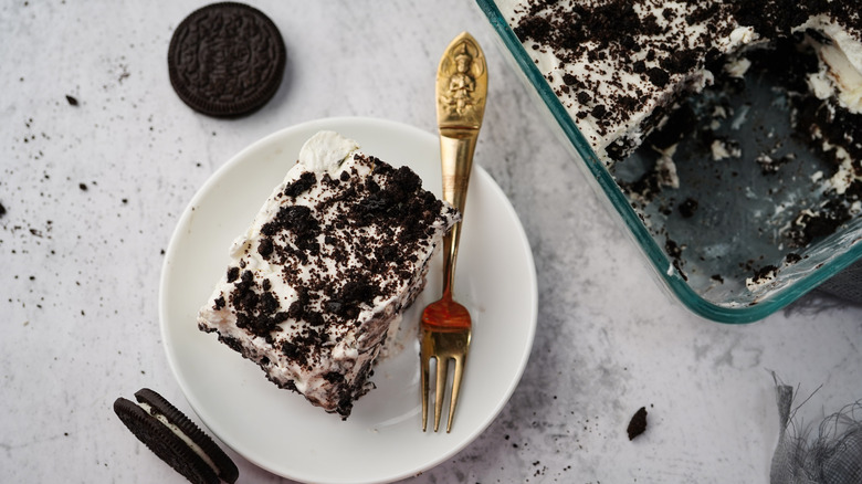 A slice of chocolate icebox cake on a plate with a gold fork next to the rest of the cake in a sheet pan