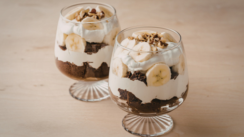 Two clear glasses filled with a dessert of pudding, cookies, banana, and whipped cream