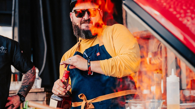 A person looking towards a flame grill at Bourbon & Beyond while holding a bottle of Maker's Mark