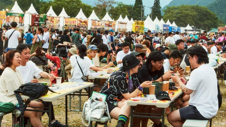 A large number of people sitting down to eat with a long row of food stands in the background