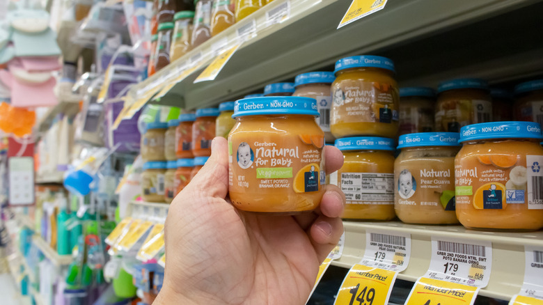 A hand holding a jar of Gerber baby food. Various other jars of Gerber baby food are pictured in the background.
