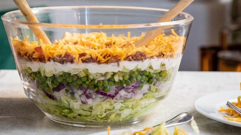 Seven layer salad in clear bowl with wooden spoons