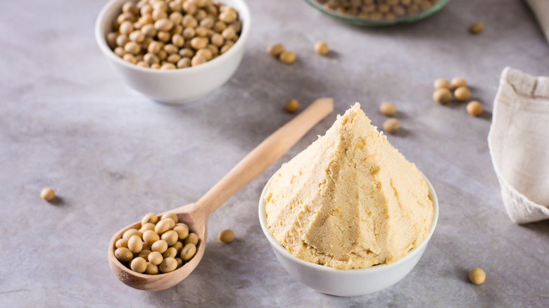 A pile of mise piso paste in a bowl surrounded by fermented soybeans on a grey background