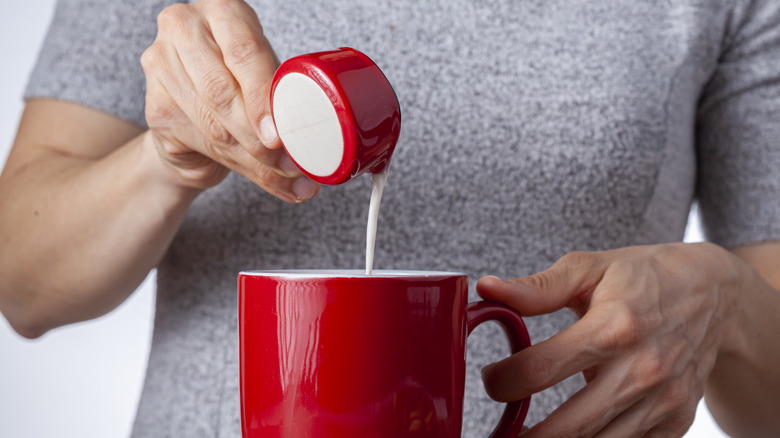 A person in a grey shirt pours creamer from a small, red creamer pitcher into a matching red mug