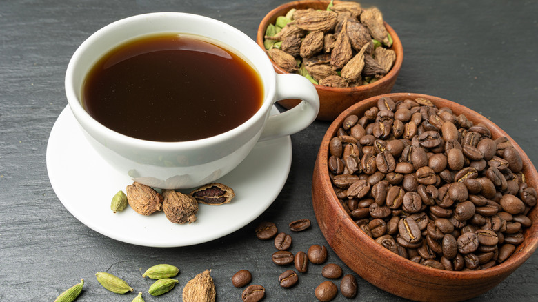 A cup of coffee next to cardamom pods and coffee beans in bowls