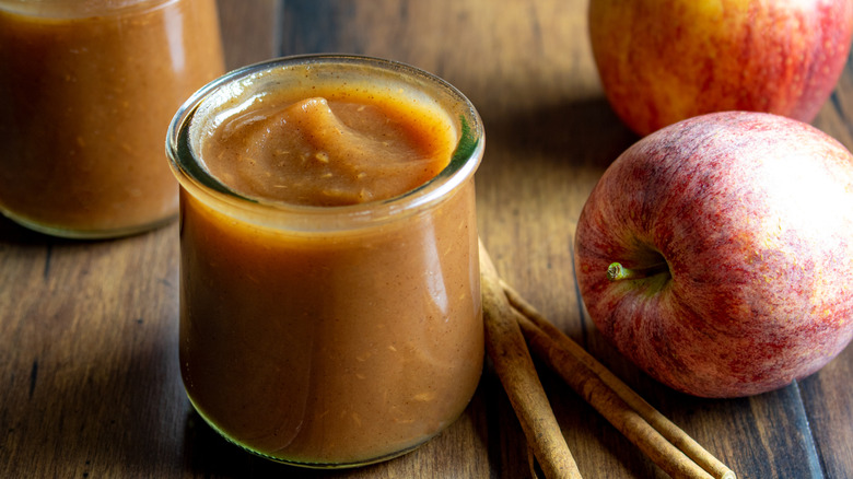 Fresh apple butter in a glass jar with whole apples and cinnamon sticks on a wood table