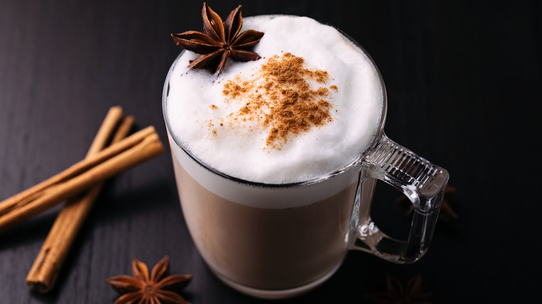A latte in a glass mug with whole spices on the side against a dark background