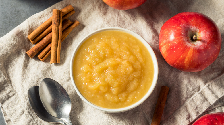 Bowl of applesauce surrounded by two spoons, apples, and cinnamon sticks