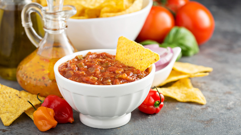 Tortilla chip dipped in bowl of salsa surrounded by ingredients and chips