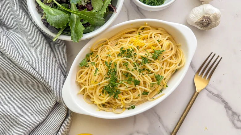 A white dish filled with spaghettini garnished with parsley next to fork and salad