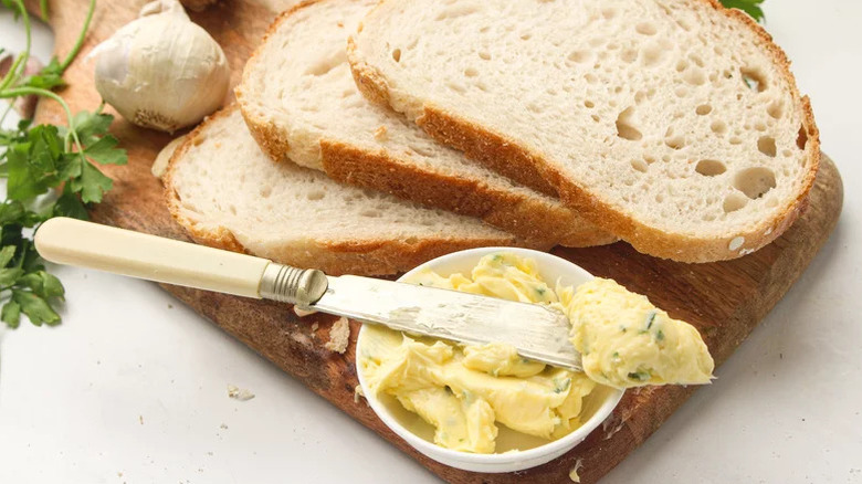 Sliced bread on chopping board with garlic butter in a dish and a butter knife
