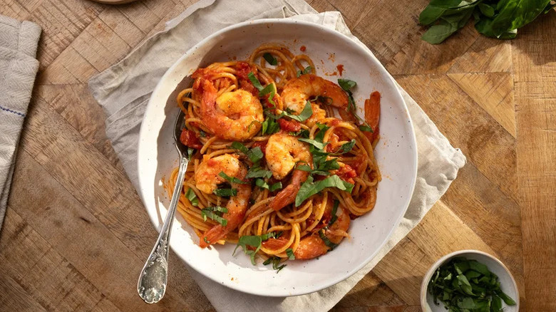 A bowl of garlic shrimp pasta with tomato sauce on a wooden table next to herbs