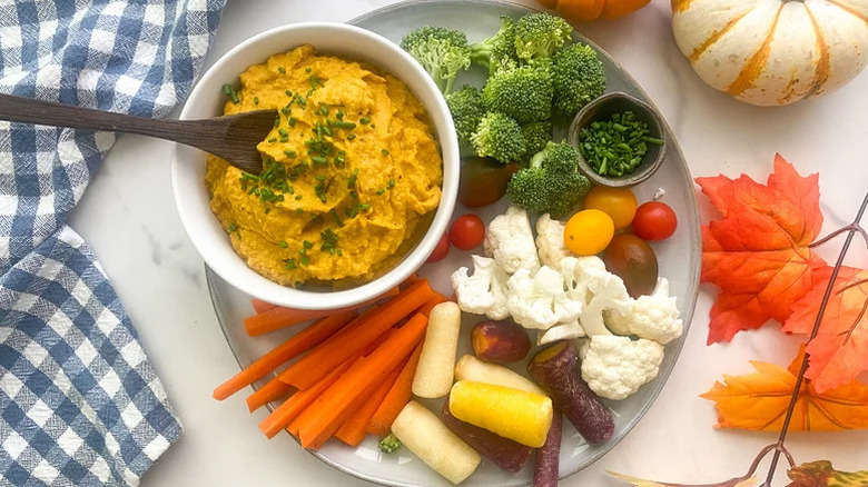 A white plate with a bowl hummus surrounded by various vegetables