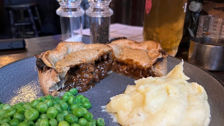 A steak and ale pie is cut in half on a plate with mashed potatoes and peas
