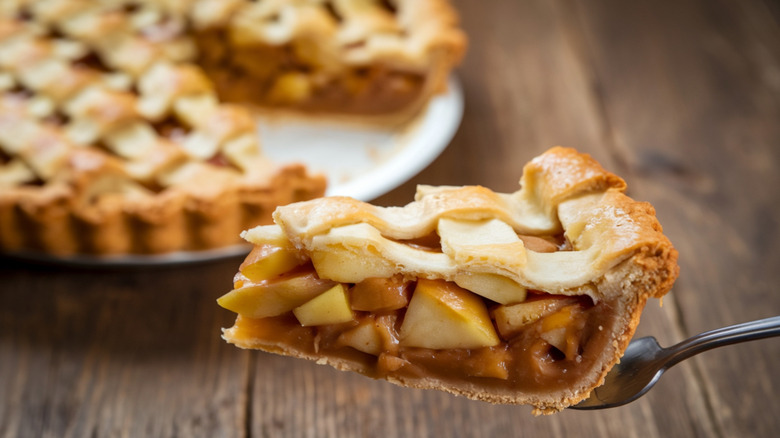 Slice of apple pie held aloft on fork, latticed pie in background