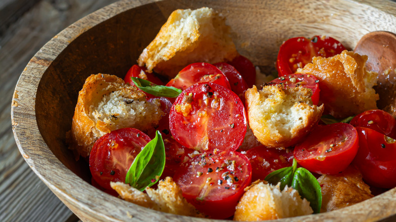 Tomatoes and chunks of bread in a wooden bowl with seasonings and fresh basil leaves