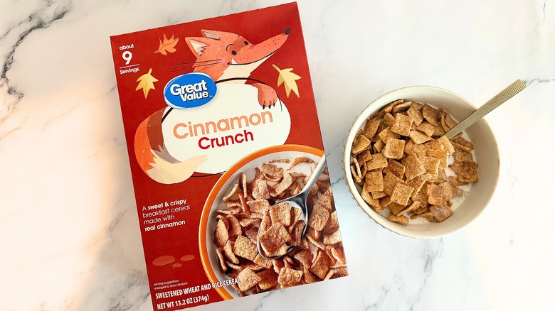 Box of Great Value Cinnamon Crunch cereal next to bowl of cereal with spoon on marble surface