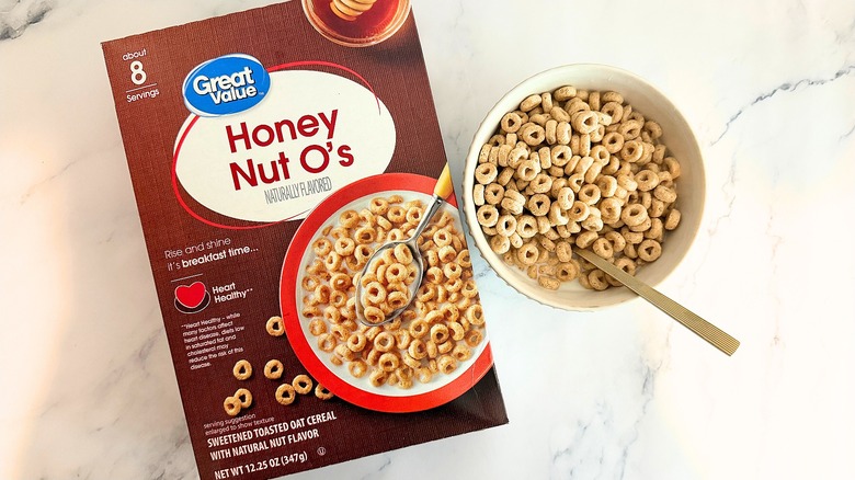 Box of Great Value Honey Nut O's cereal next to bowl of cereal with spoon on marble surface