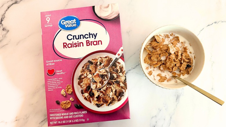Box of Great Value Crunchy Raisin Bran cereal next to bowl of cereal with spoon on marble surface