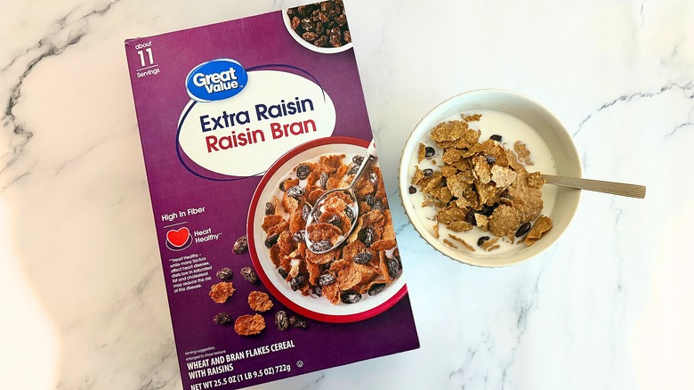 Box of Great Value Extra Raisin Raisin Bran cereal next to bowl of cereal with spoon on marble surface