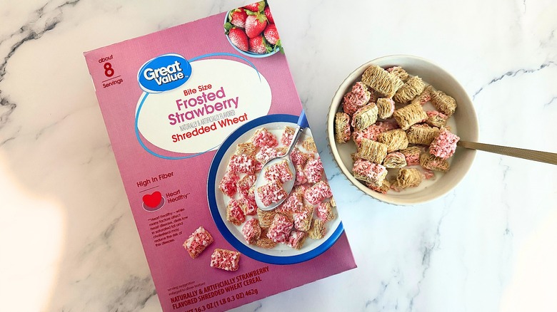 Box of Great Value Bite Size Frosted Strawberry Shredded Wheat cereal next to bowl of cereal with spoon on marble surface