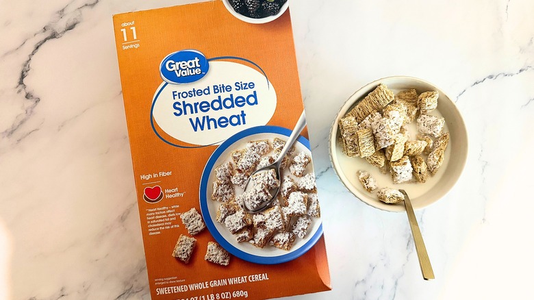 Box of Great Value Frosted Bite Size Shredded Wheat cereal next to bowl of cereal with spoon on marble surface