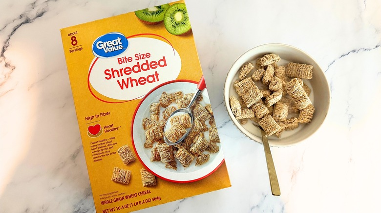 Box of Great Value Bite Size Shredded Wheat cereal next to bowl of cereal with spoon on marble surface