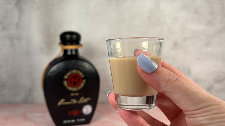 Hand holding shot glass of cream liqueur in front of blurry black bottle of liqueur