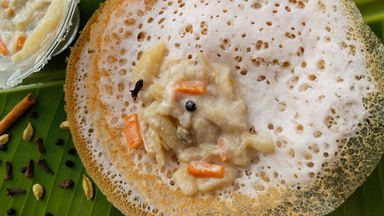 Appam topped with vegetable stew, served on banana leaf sprinkled with spices